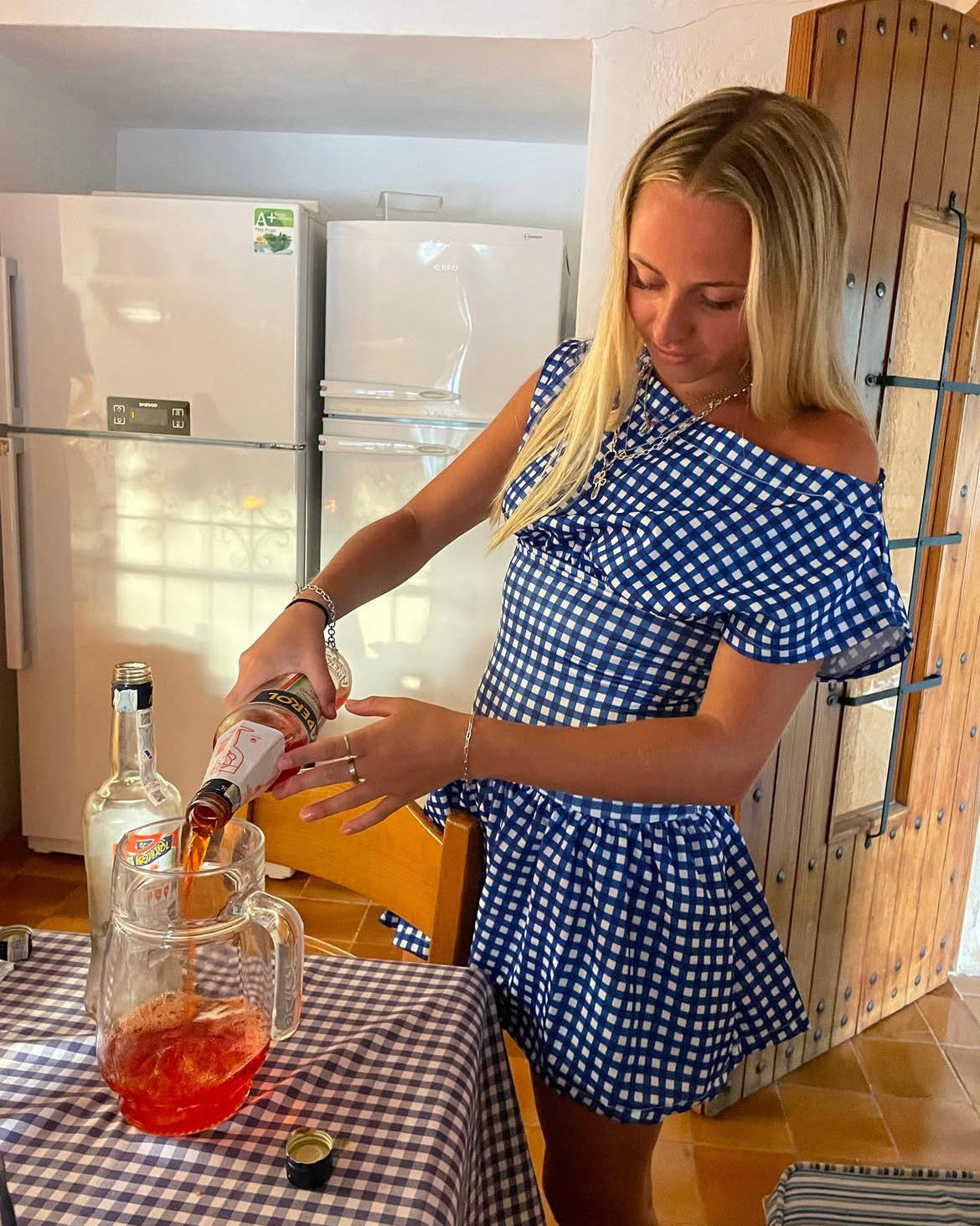Woman in a blue checkered dress pouring a drink into a glass at a table.