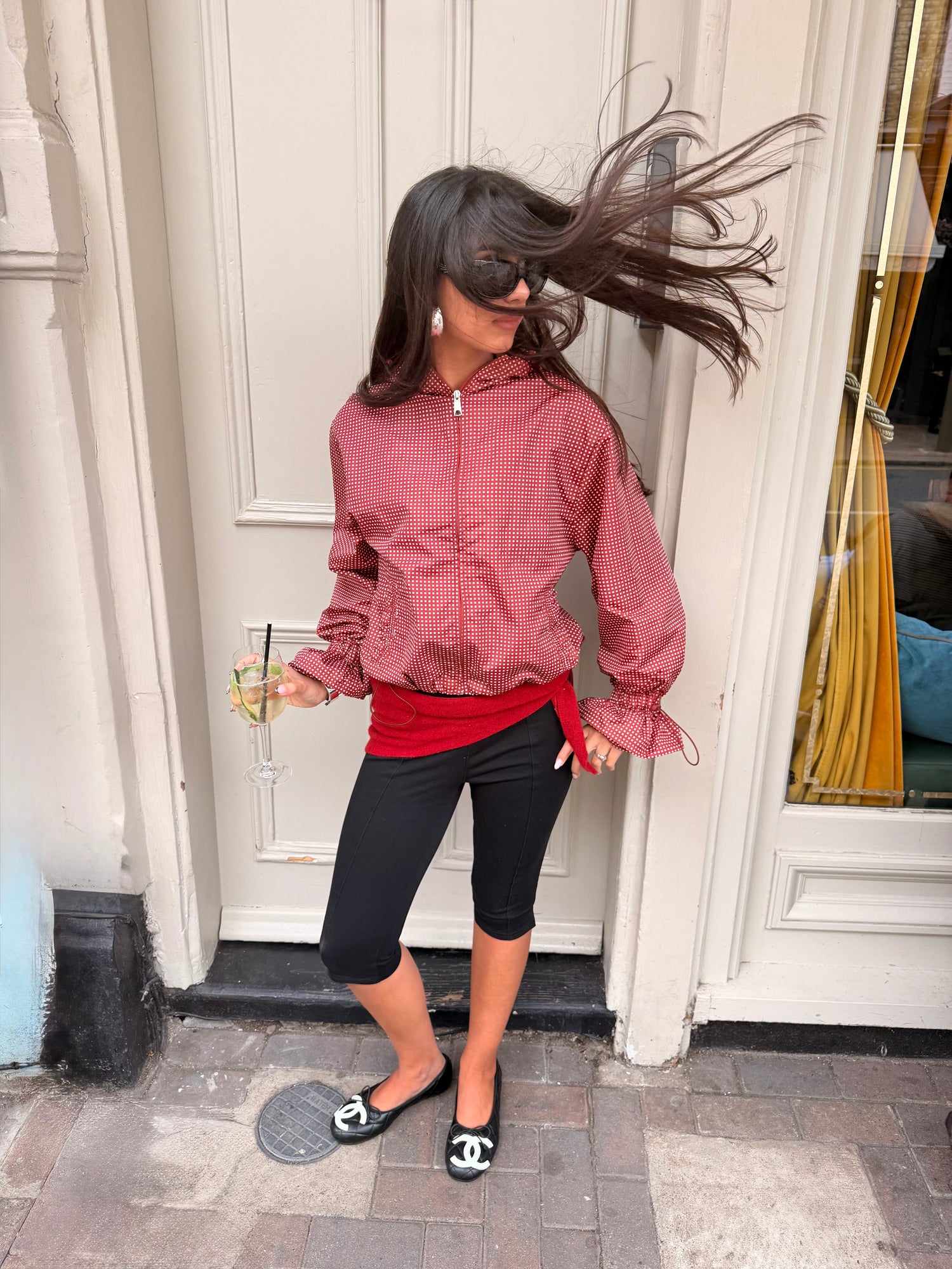 A woman posing outdoors in front of a white door, wearing a dusty rose zip-up jacket over a bright red top, black capri leggings, and black-and-white Mary Jane shoes.