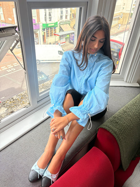 A woman sits on a window ledge wearing a light blue and white gingham long-sleeve blouse with dramatic puffed sleeves, paired with black bottoms and light blue-and-black flats. The background shows a street scene visible through the window.