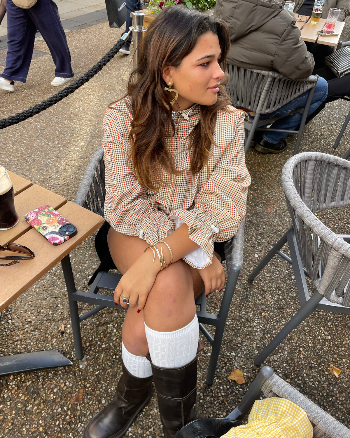 A woman seated outdoors wearing a beige and brown plaid windbreaker, white long sleeves, and black boots.