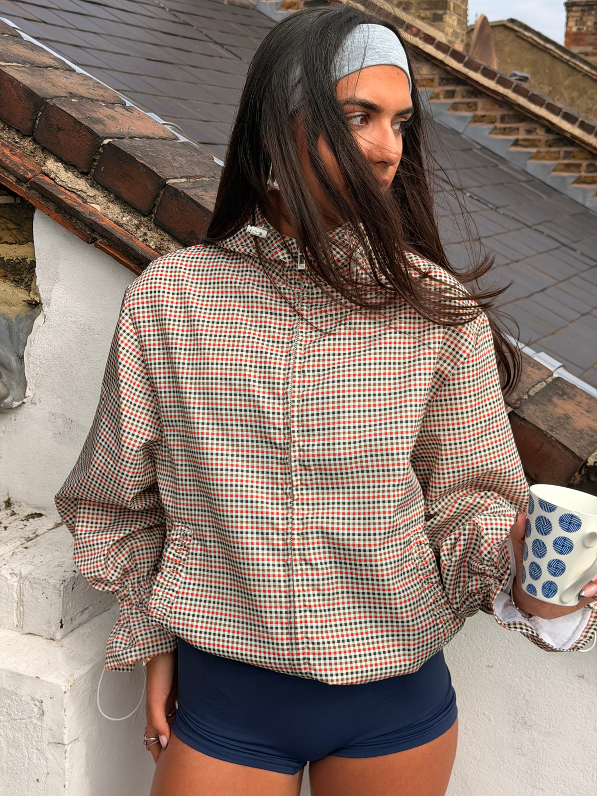 A woman wearing a beige and red gingham long-sleeve button-up shirt and navy blue shorts, holding a blue and white polka dot mug against a background of brick and tiled rooftops.