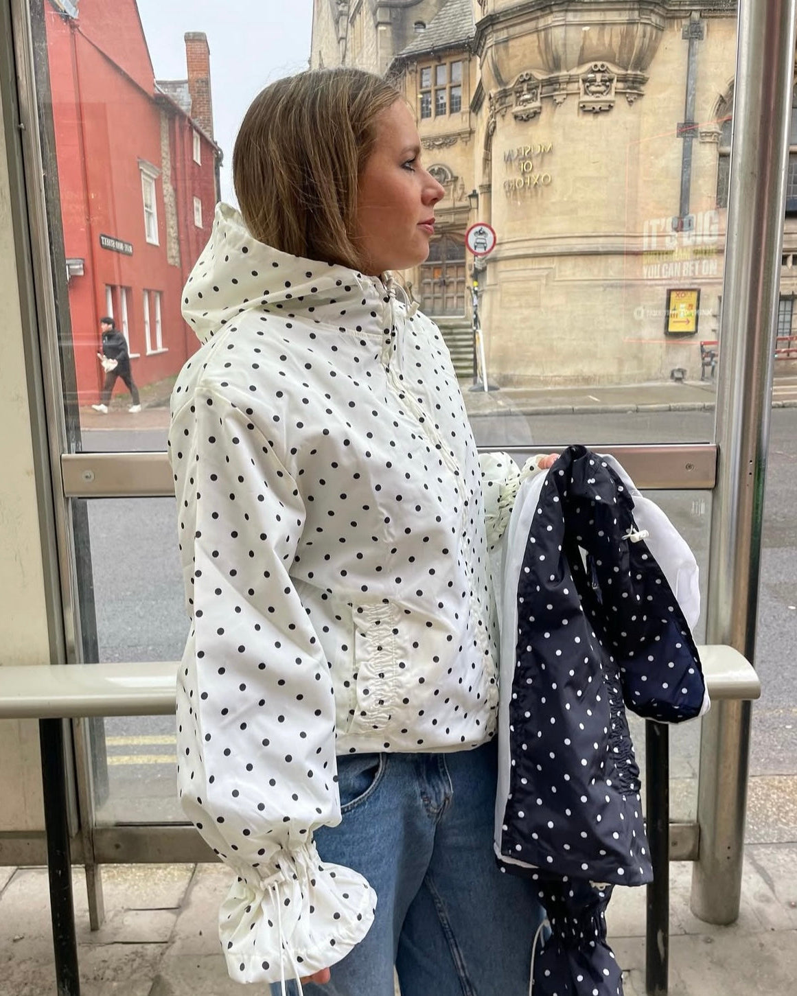 Person wearing a white raincoat with black polka dots at a bus stop.