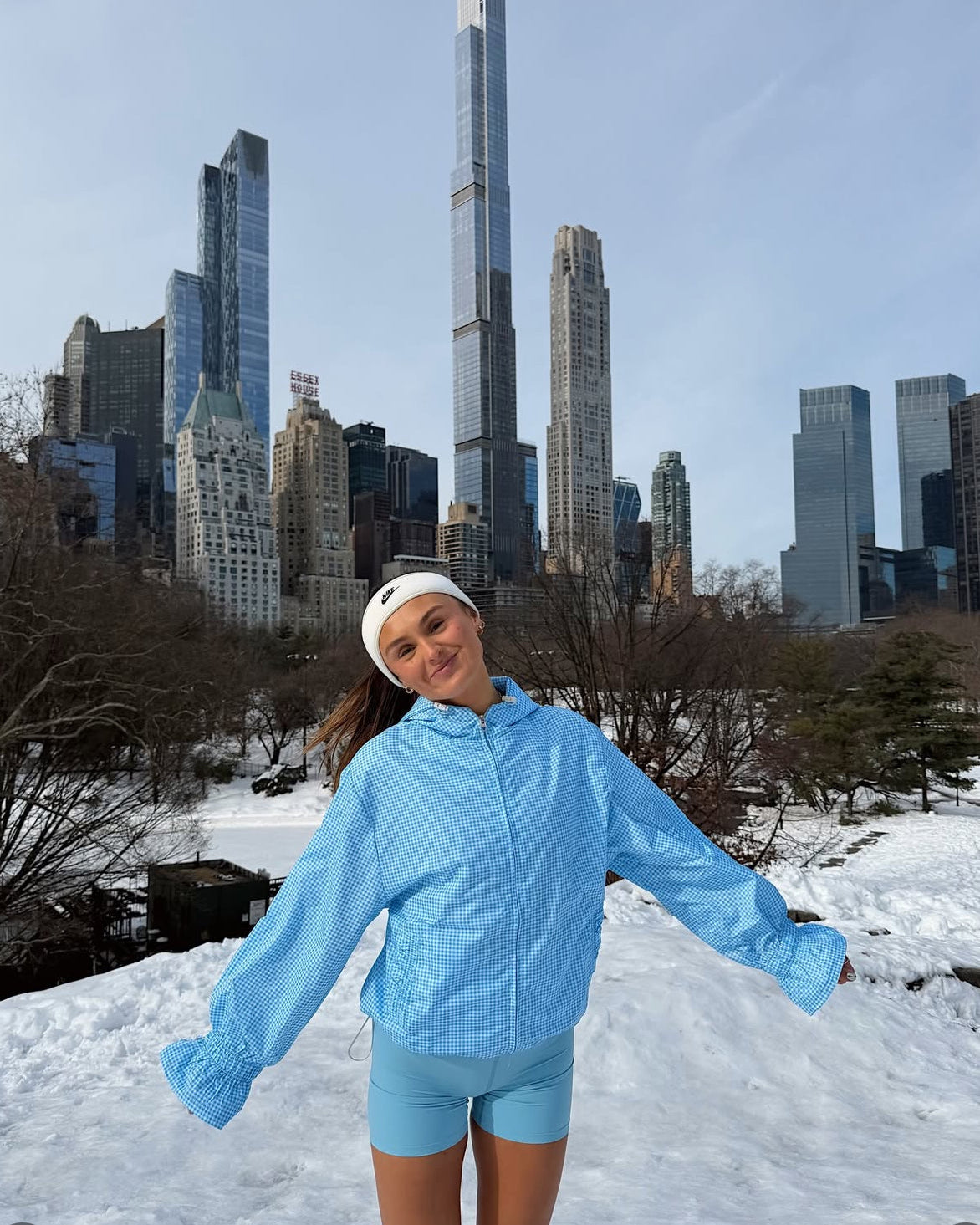 Person in a blue jacket and shorts standing in a snowy park with city skyline in the background