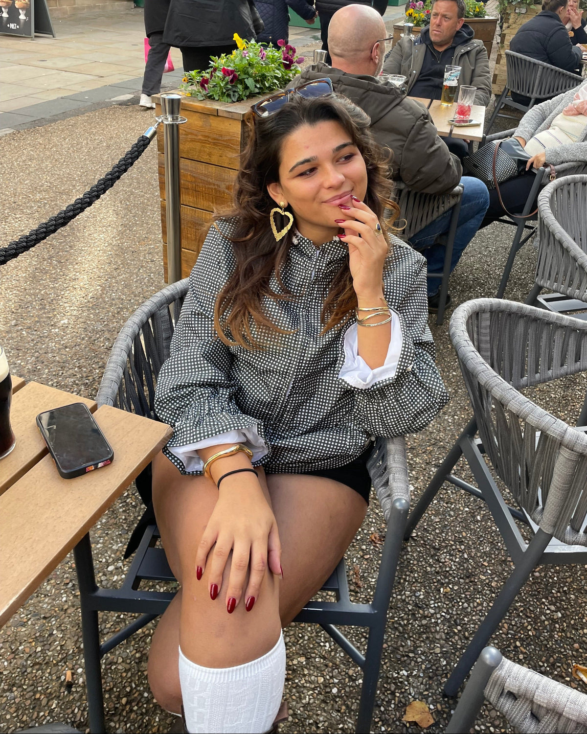 A woman sitting at an outdoor cafe table wearing olive gingham patterned zip-up jacket over a white collared shirt, paired with black shorts and white socks.