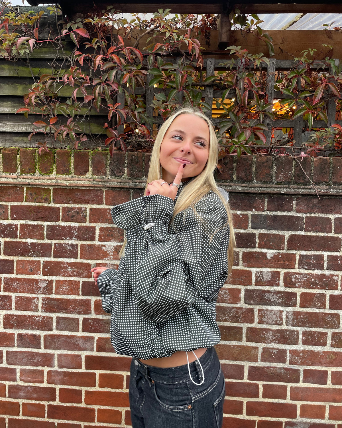 A blonde woman posing in front of a brick wall and leafy vines, wearing a olive gingham cropped jacket and dark jeans.