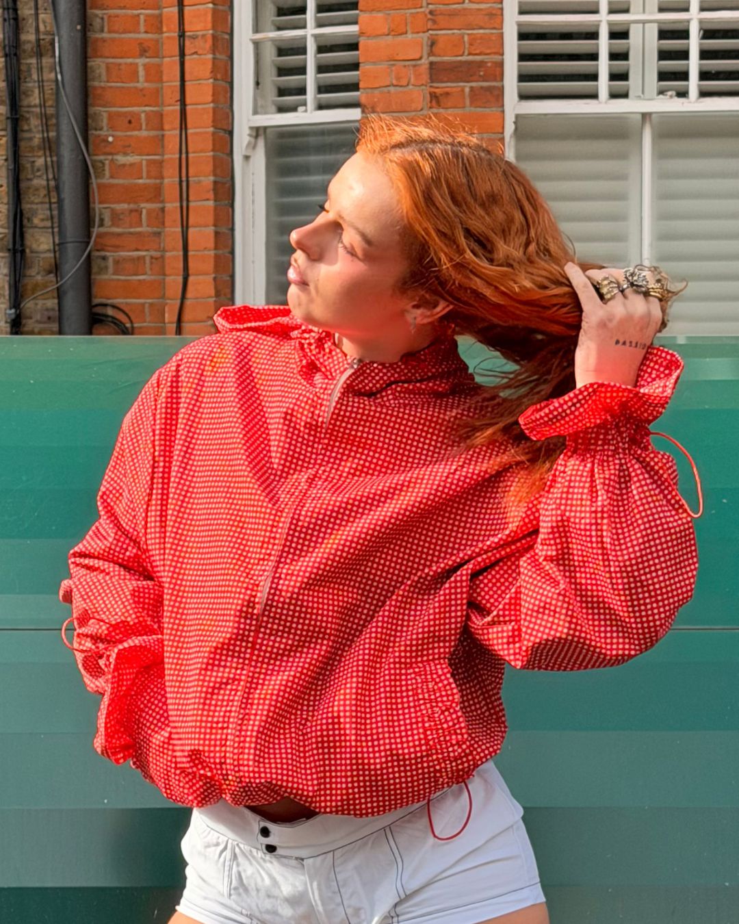 A woman with red hair wearing a red and white gingham patterned long-sleeve shirt and white shorts, posing outdoors in front of a green wall and brick building.