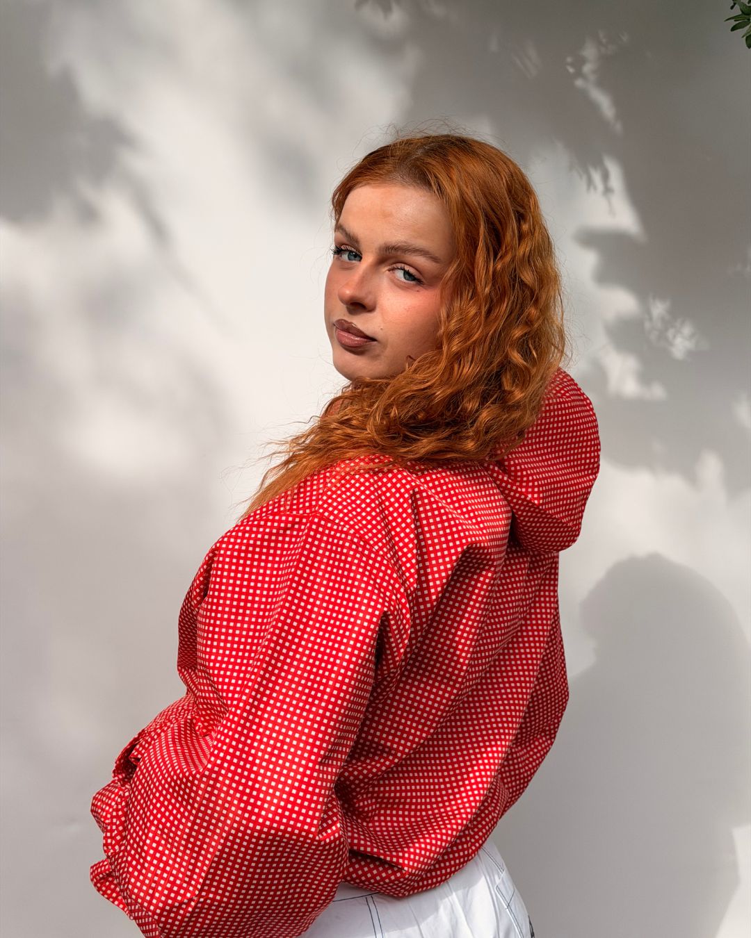 A woman with curly red hair wearing a red and white polka dot hooded jacket, posing against a white wall with soft leaf shadows.