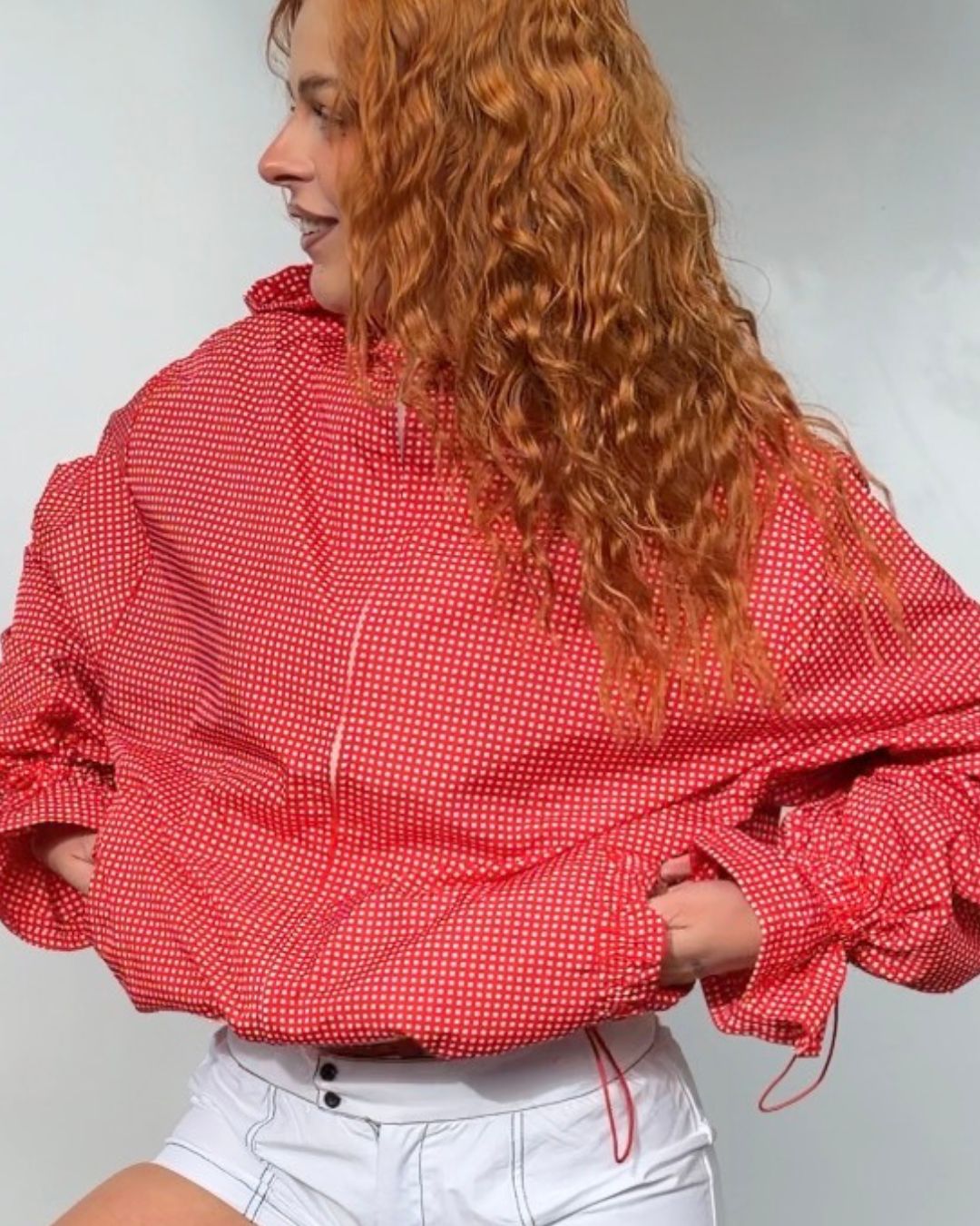 A woman with long curly red hair wearing a red and white gingham patterned oversized zip-up jacket and white shorts, shown from the side against a light gray background.