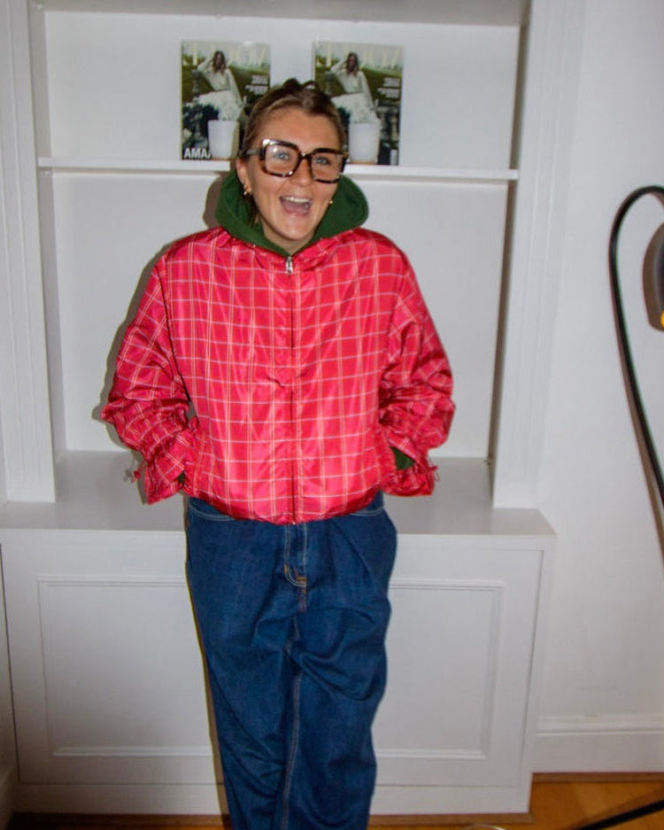 Person wearing a red plaid jacket and blue pants standing in a room with a white bookshelf and lamp.