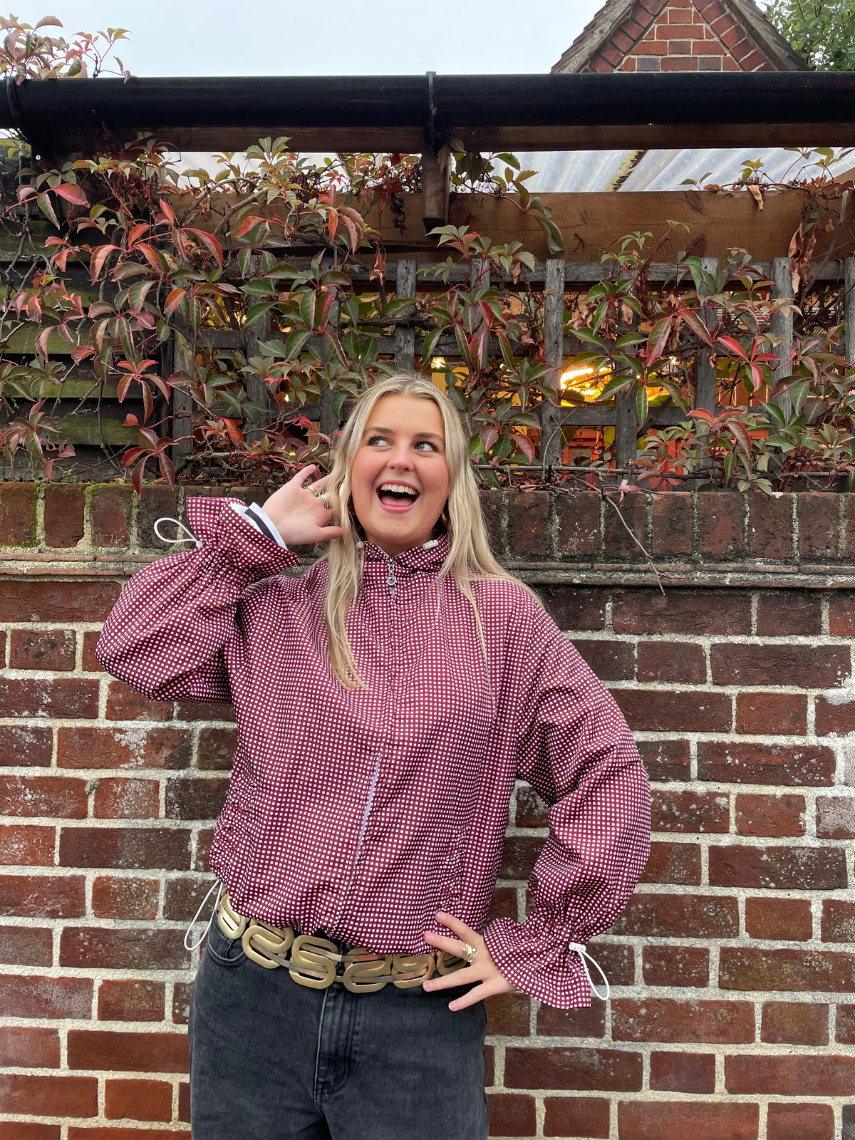 a woman against a brick wall wearing a cherry gingham windbreaker jacket with flared sleeves styled with jeans
