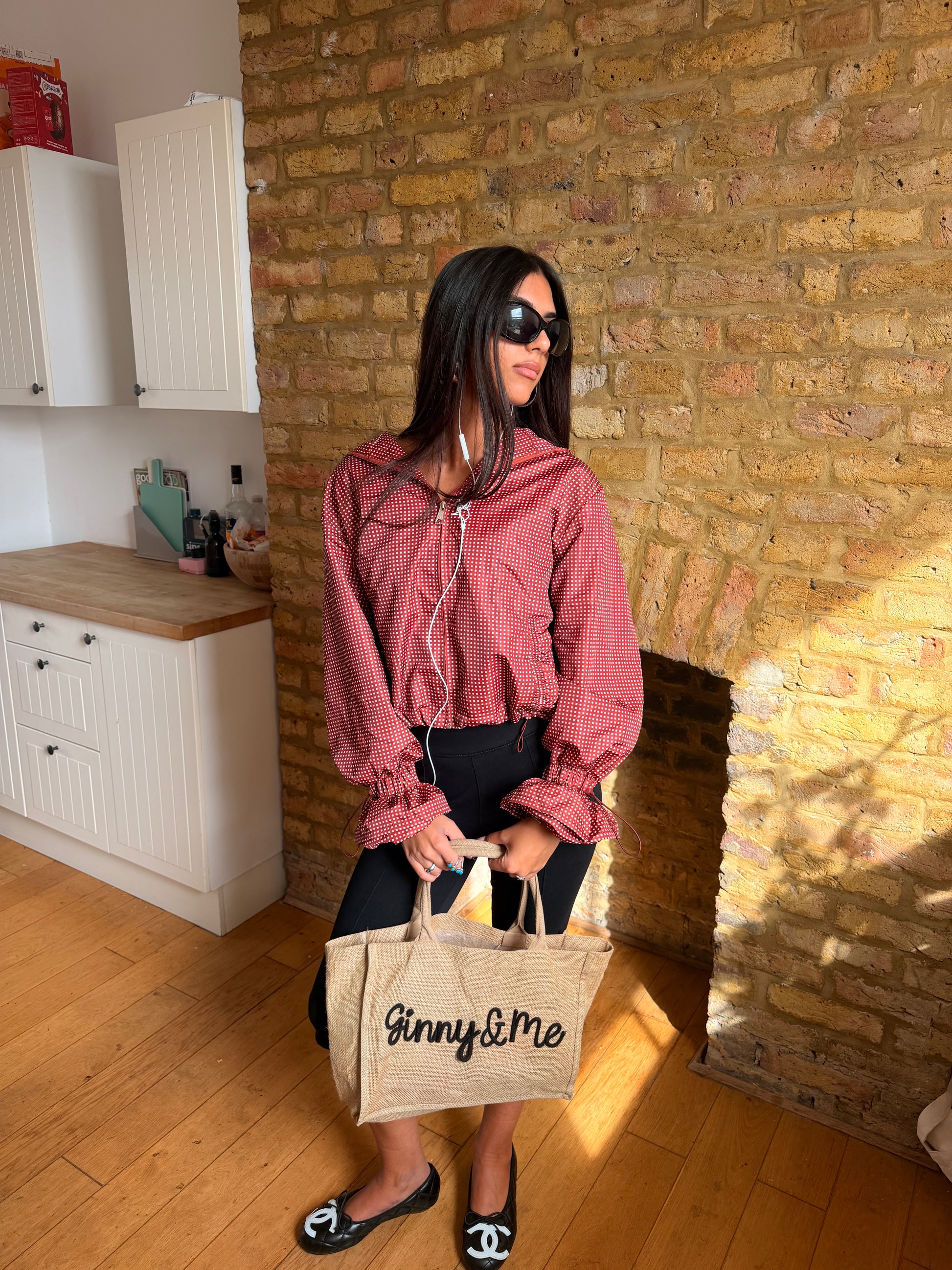 A woman stands in a room with a brick wall and wooden floor, wearing a dusty rose cropped jacket, black pants, and black flats with a Chanel logo. She is holding a beige tote bag with 'Ginny & Me' printed on it.