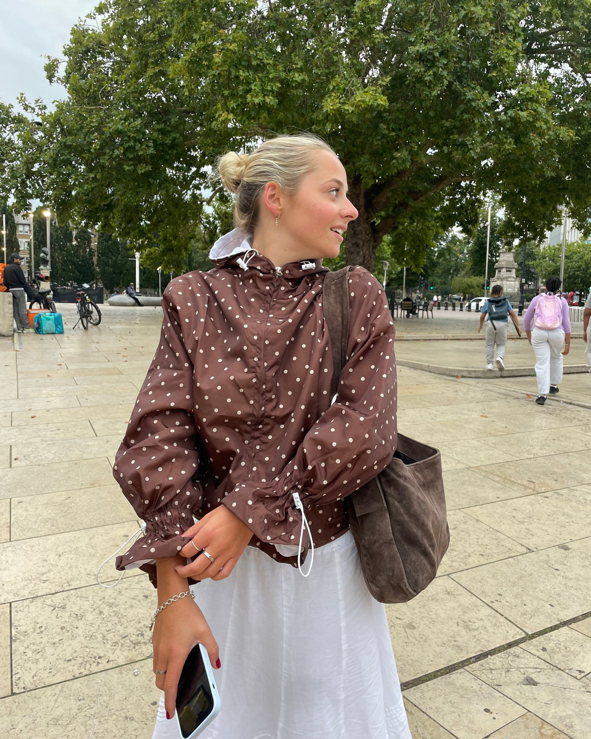 Person in a brown polka dot coat standing on a sidewalk with trees and people in the background.