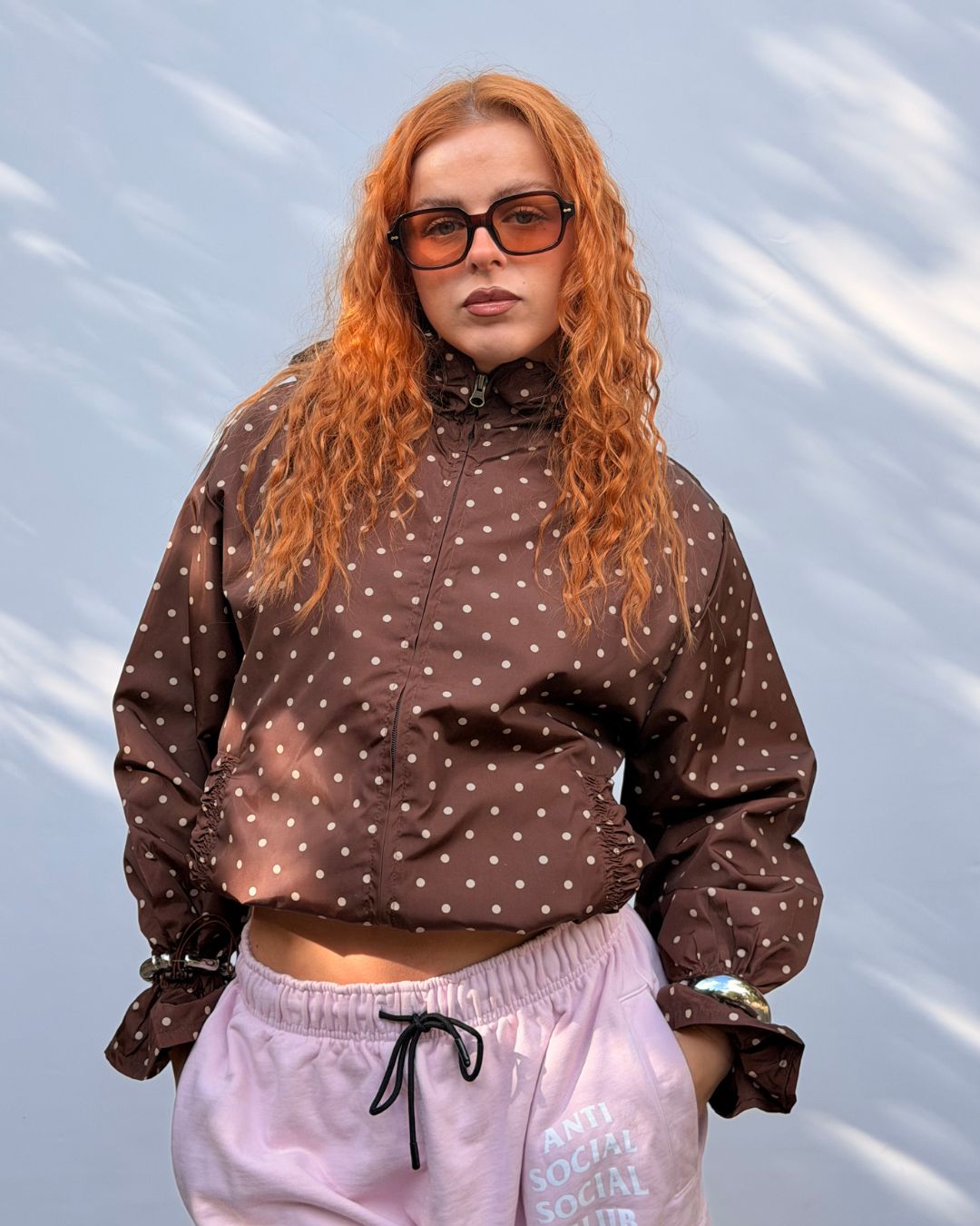 A woman with long curly red hair and black sunglasses wearing a brown cropped puffer jacket with white polka dots and light pink sweatpants, standing against a light blue textured background.