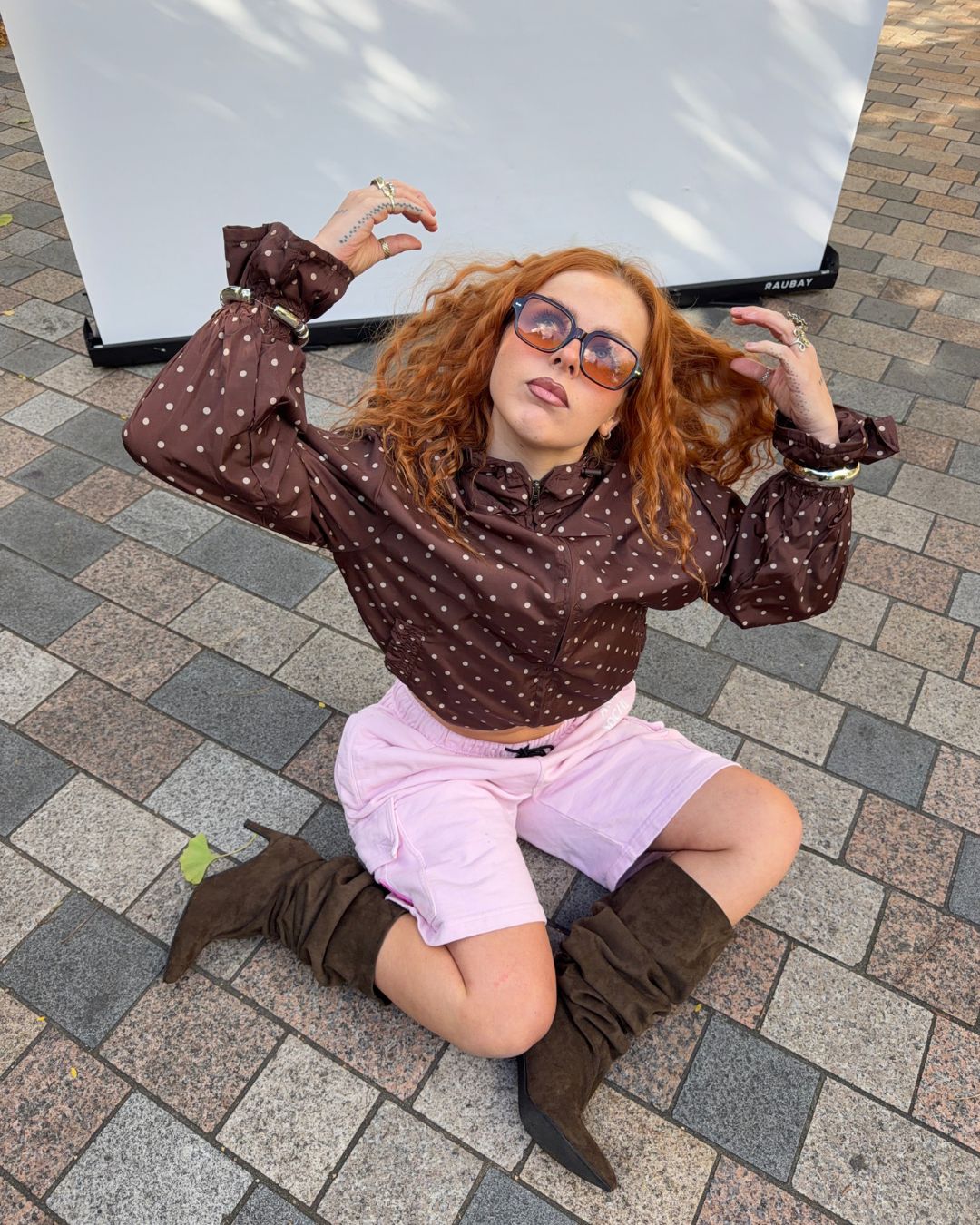 A woman with red hair wearing a brown long-sleeve button-up shirt with white polka dots, light pink shorts, and brown knee-high boots, sitting on a paved outdoor surface in front of a white screen.