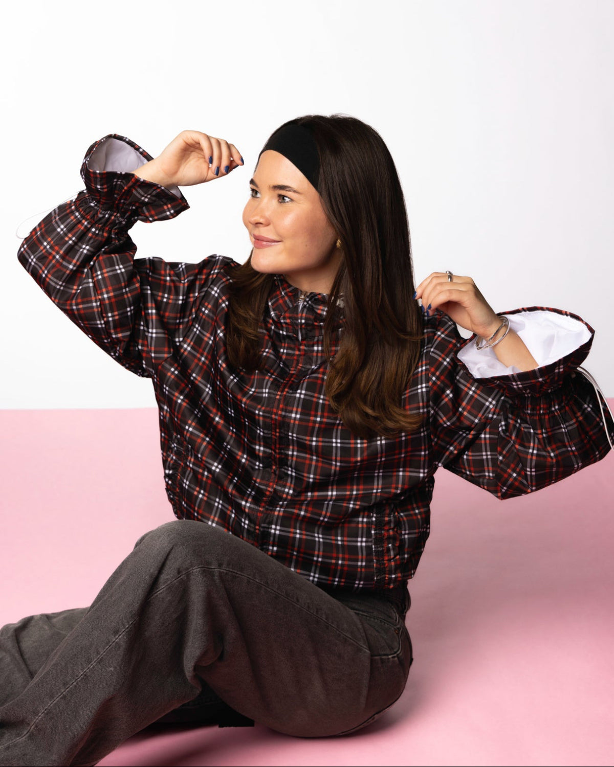 Person wearing a plaid windbreaker sitting on a pink surface with a white background