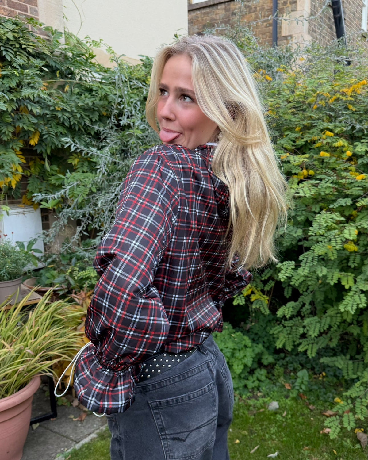 Woman in a plaid shirt and jeans standing outdoors with greenery and a building in the background