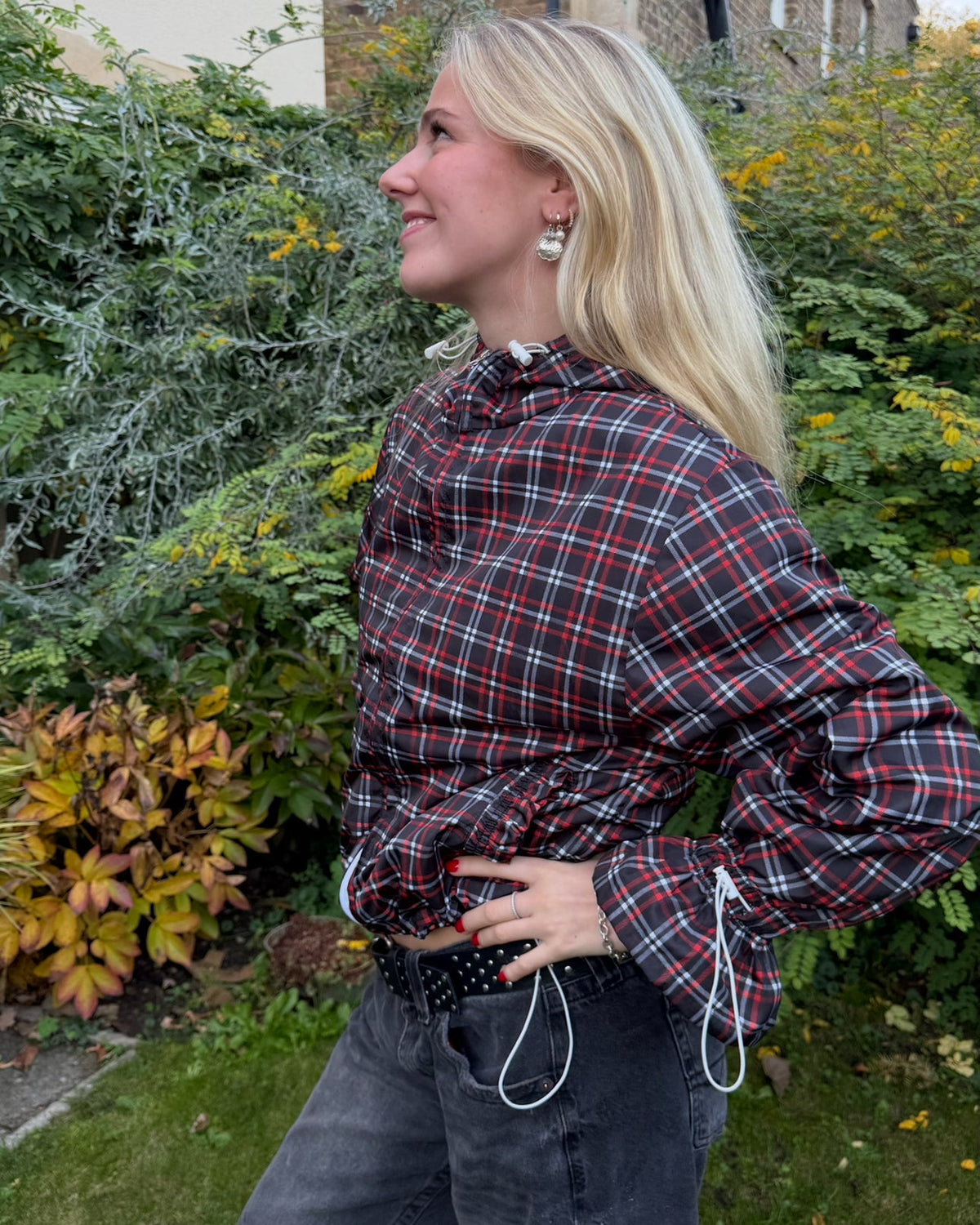 Person wearing a plaid windbreaker and jeans standing outdoors with greenery and a building in the background