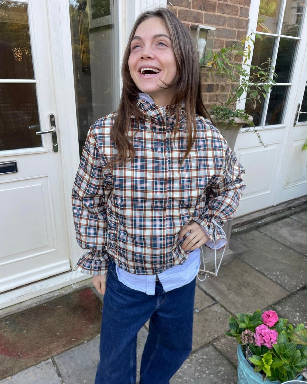 Person wearing a plaid shirt and blue pants standing outside a house.