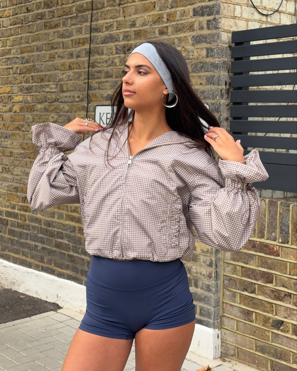 A woman posing outdoors against a brick wall, wearing a light brown and white gingham cropped zip-up hoodie with dramatic balloon sleeves, paired with navy blue athletic shorts.