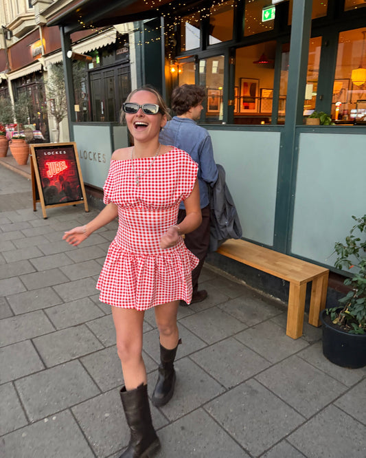 Person wearing a red checkered dress standing on a sidewalk with a building in the background