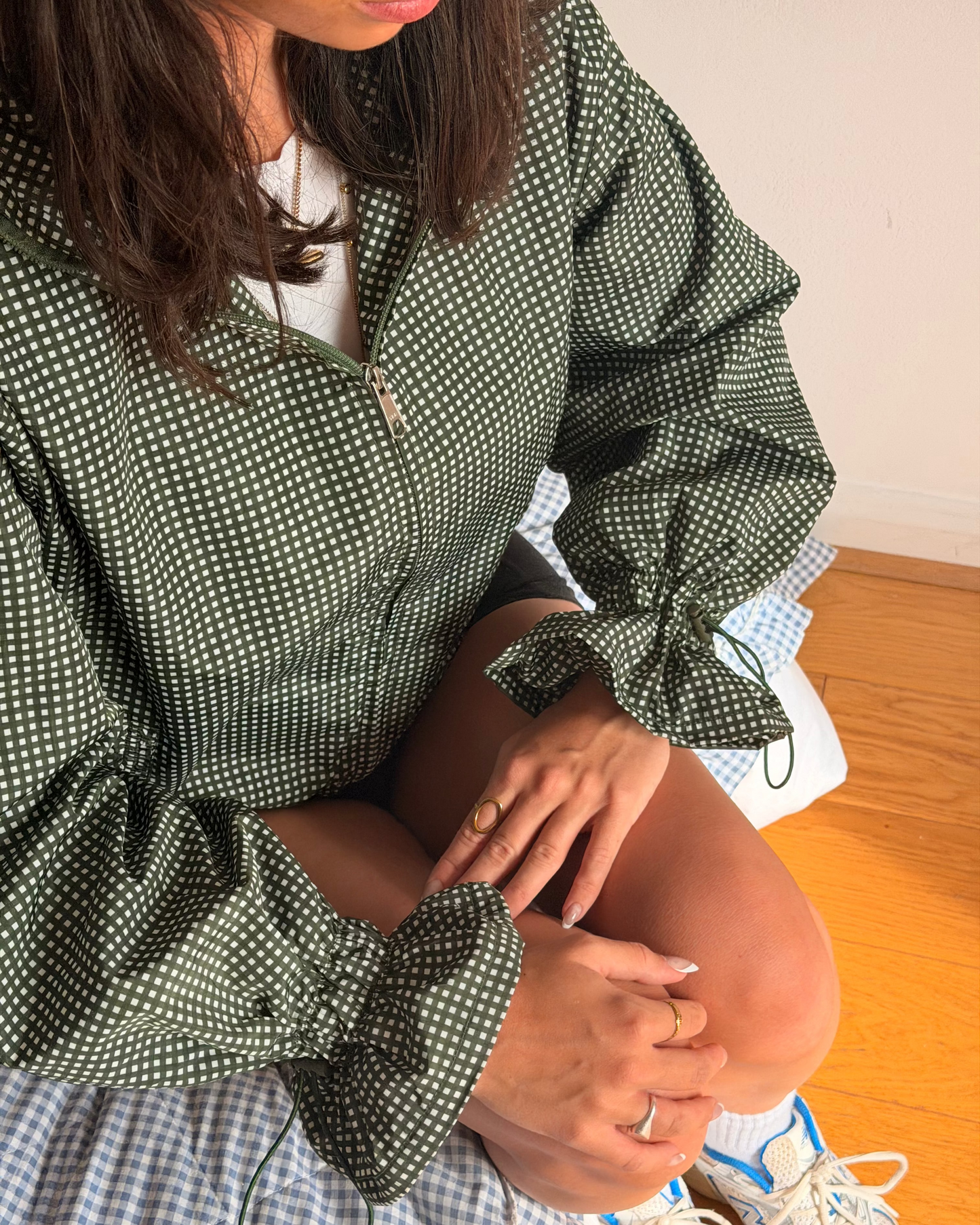 A woman wearing a green and white gingham patterned zip-up jacket with tie details on the sleeves, paired with a blue and white gingham skirt and white sneakers.