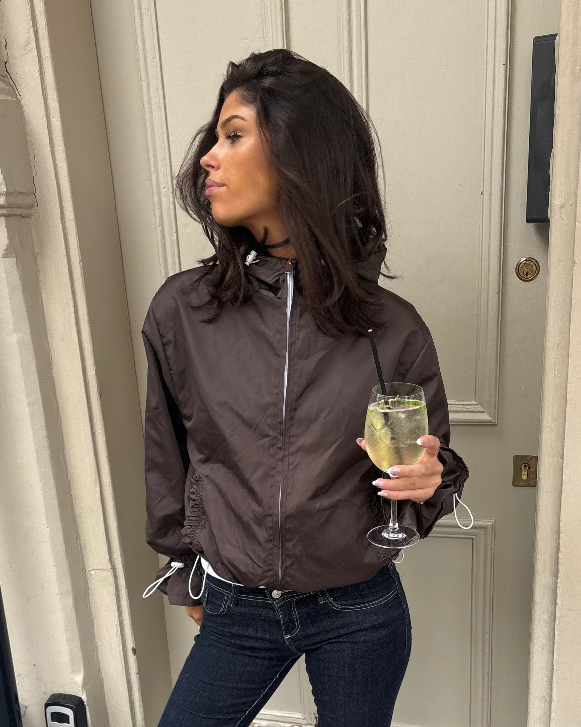 A woman wearing a dark brown lightweight zip-up jacket, dark blue flared jeans, and tan suede boots stands in front of a cream-colored door, holding a glass of champagne.