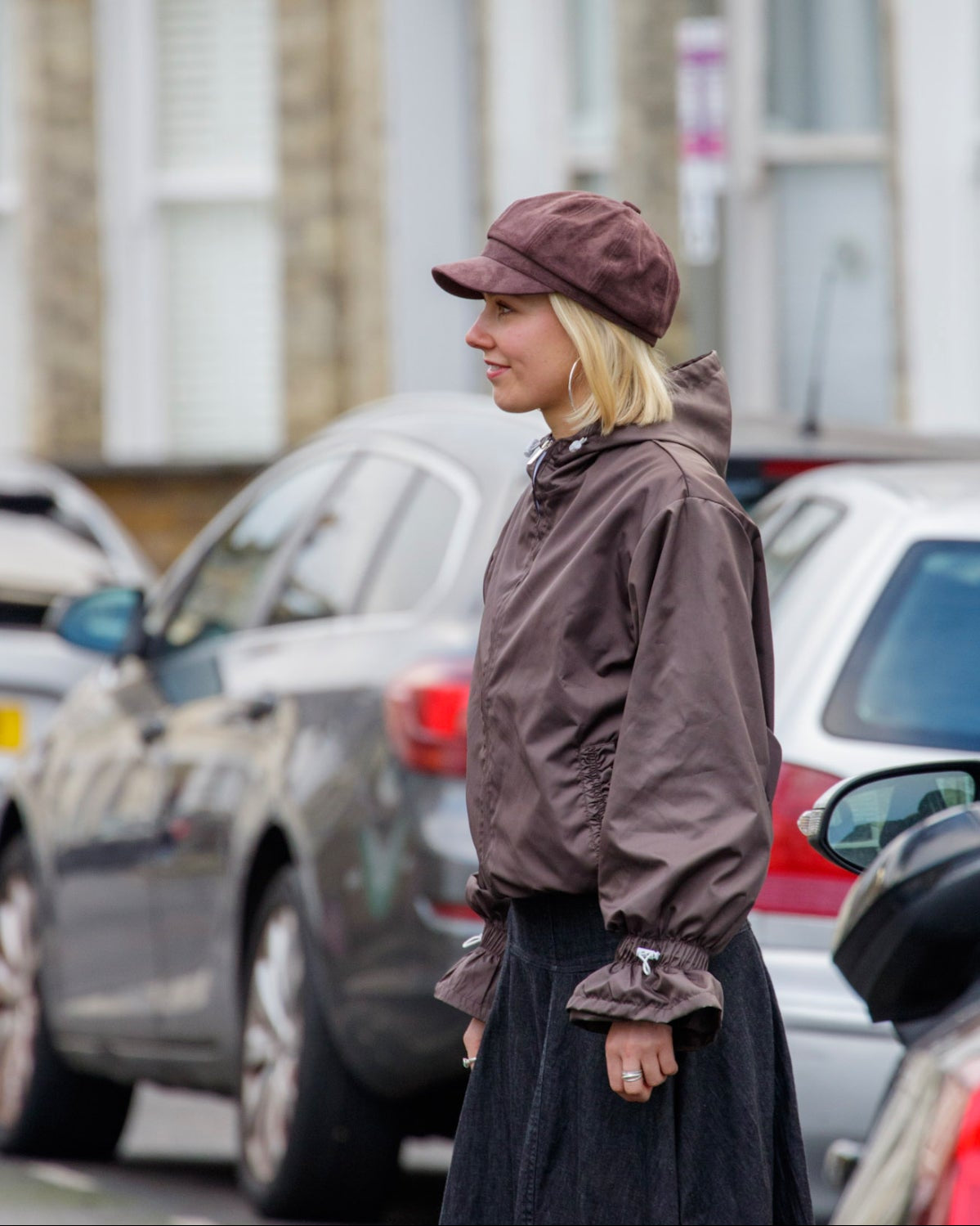 Woman in a brown jacket and cap standing on a street with cars and buildings in the background