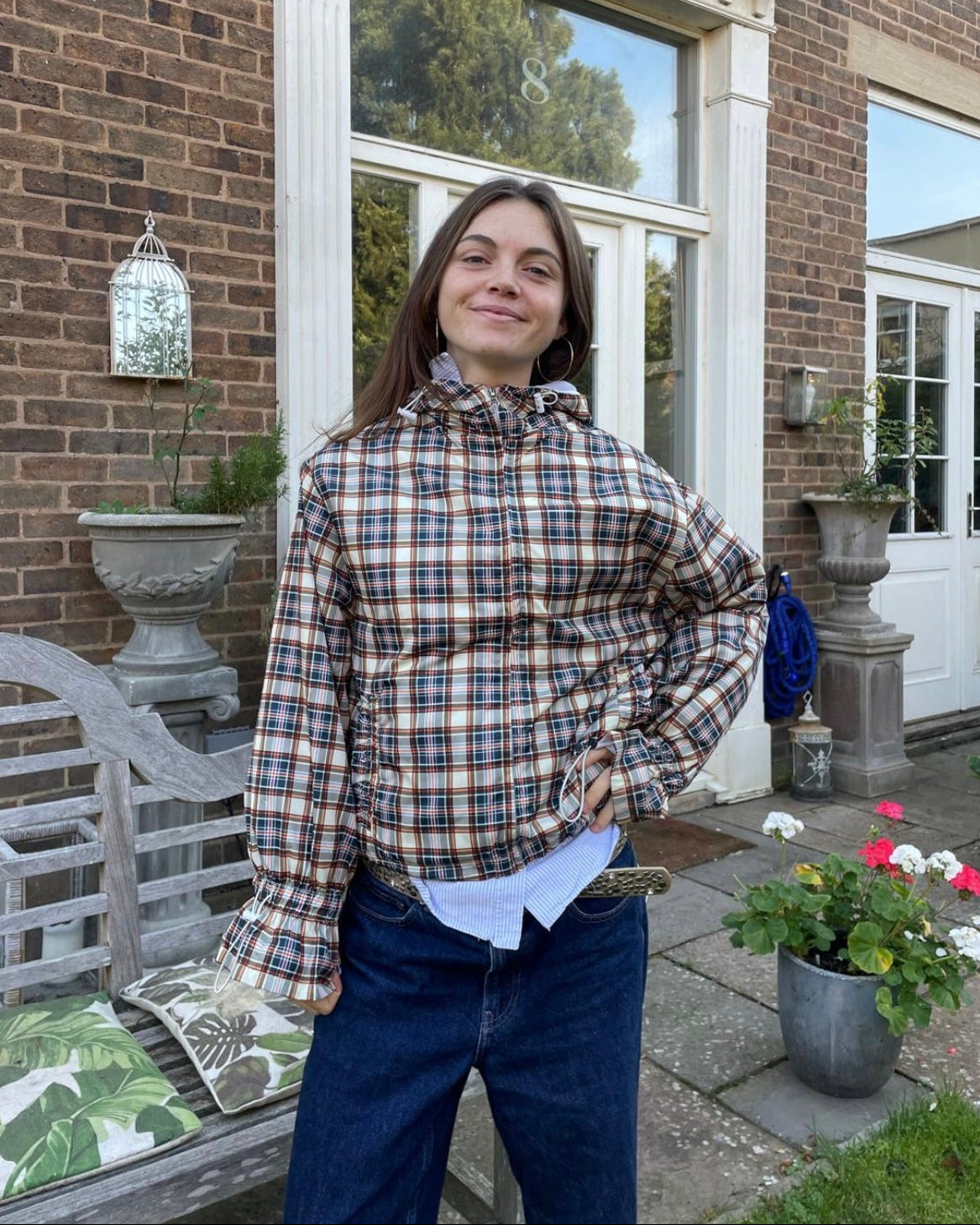 Person standing on a porch with a plaid shirt and blue jeans, surrounded by potted plants and a brick house.