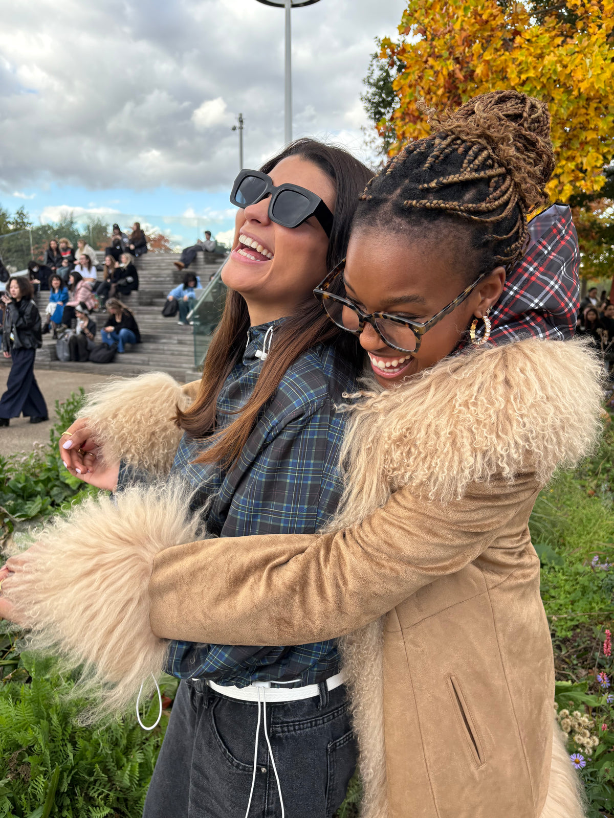 Two women hugging outdoors with a cloudy sky and trees in the background