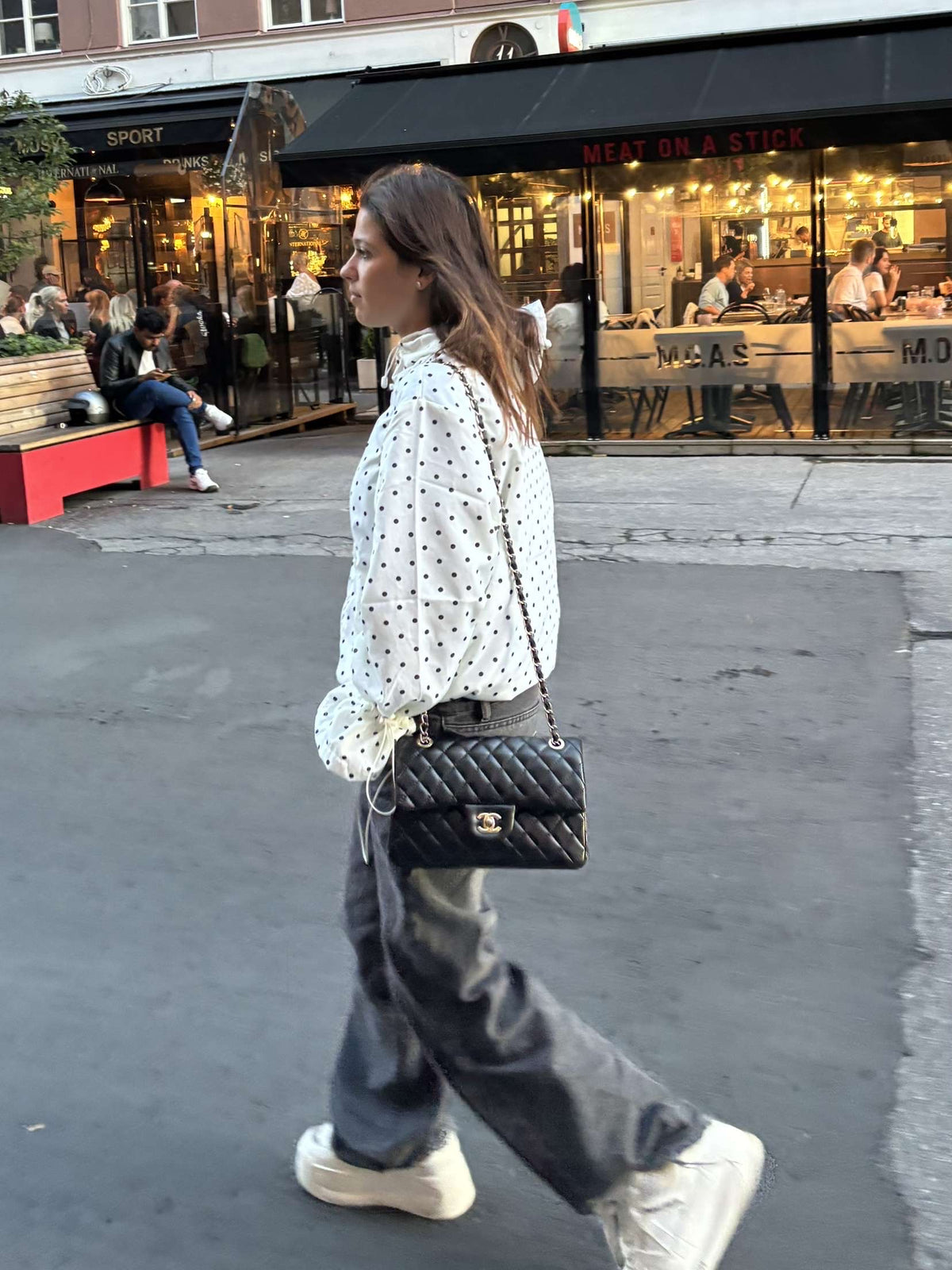 Woman walking on a street with a black handbag, wearing a white polka dot shirt and gray pants.
