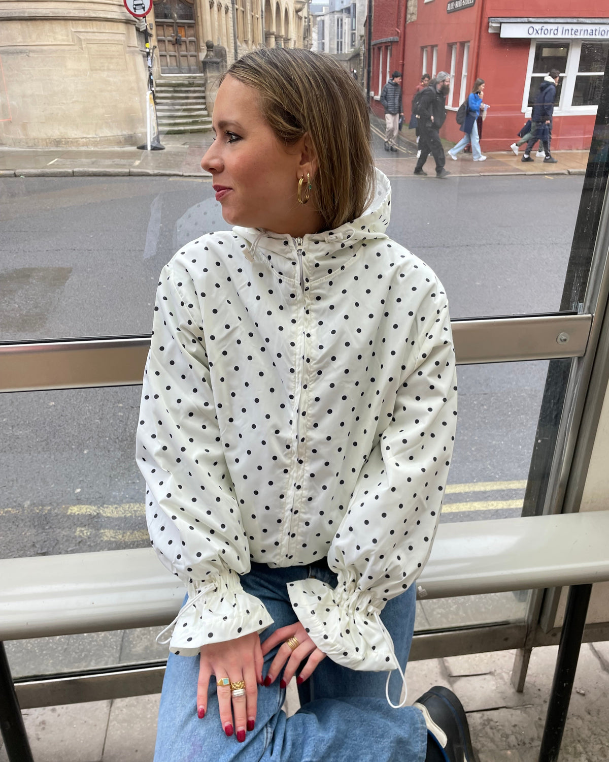 A person standing on the side of a road wearing a white and navy polka dot waterproof windbreaker with a hood, paired with blue jeans.