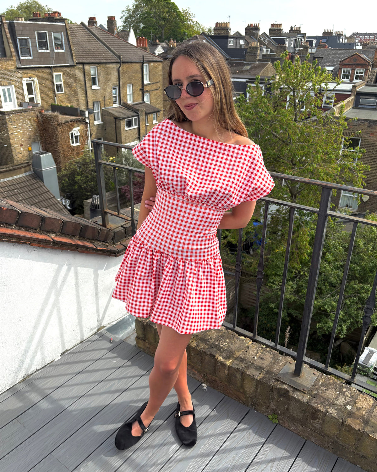 Woman in a red and white checkered dress standing on a rooftop with houses and trees in the background.
