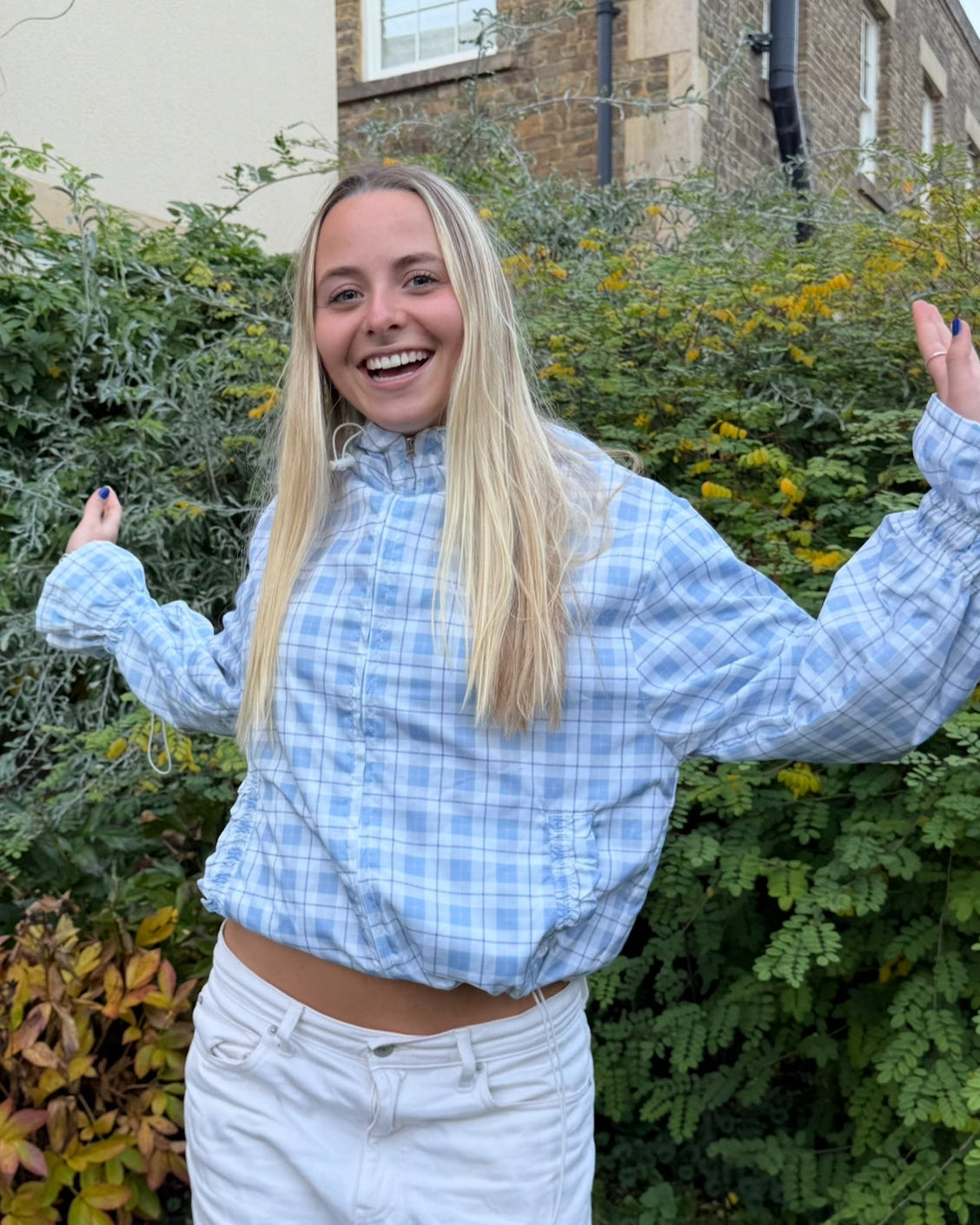Person wearing a blue checkered shirt and white pants standing outdoors with greenery and a building in the background.