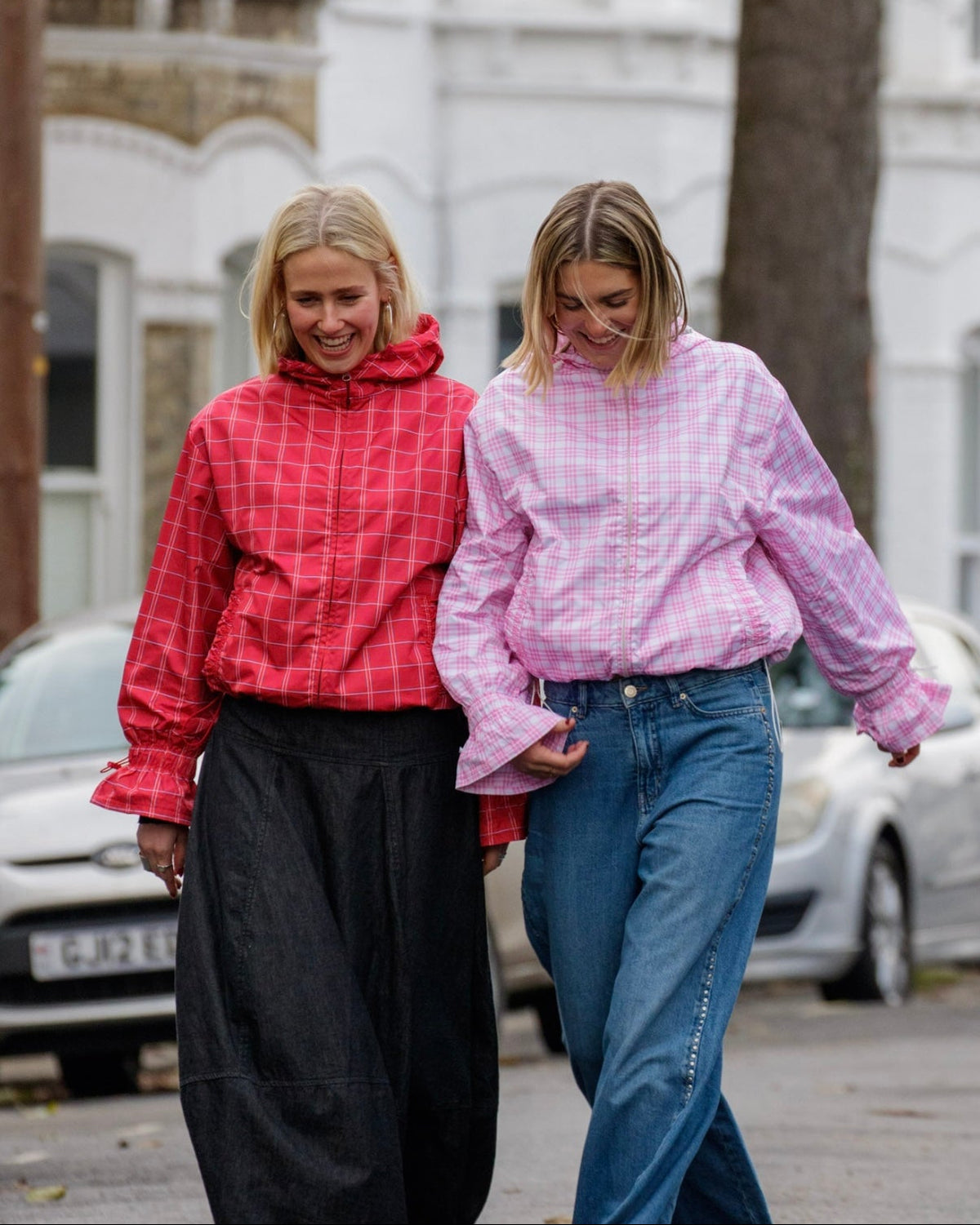 Two women walking together on a street, one in a red checkered jacket and black pants, the other in a pink checkered jacket and blue jeans.