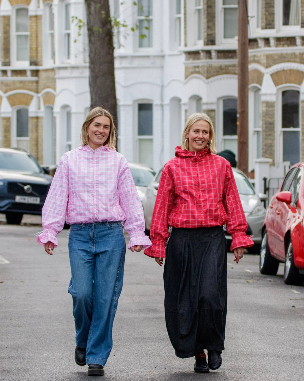 Two women walking on a street, one in a pink checkered shirt and blue jeans, the other in a red plaid shirt and black pants.